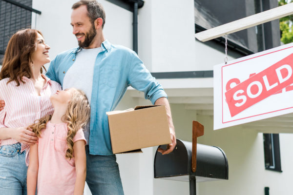 panoramic shot of happy man holding box and standing with family near house and board with sold letters